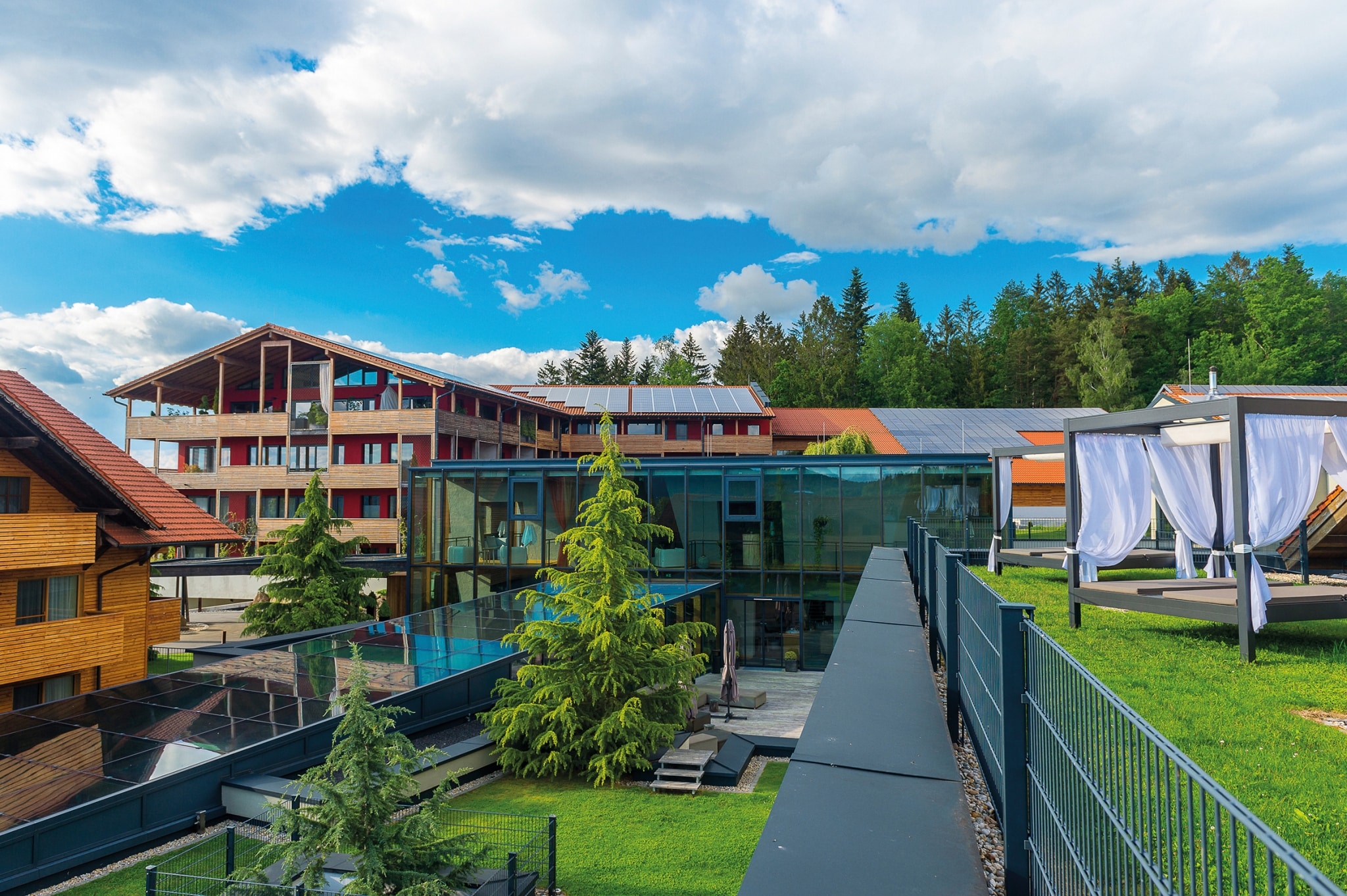 Außenansicht des ULRICHSHOFs an einem sonnigen Tag, mit modernem Glasbau und traditionellen Holzelementen. Im Vordergrund sind stilvolle Außenbereiche mit Liegen und überdachten Betten zu sehen, während im Hintergrund der Hauptgebäudekomplex mit Solarpaneelen auf dem Dach vor einem Wald liegt.