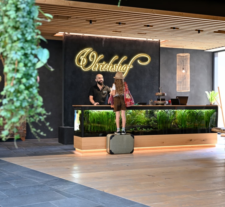 Ein Mädchen steht auf einem Koffer vor einem Aquarium im ULRICHSHOF Lobby Foyer. Dahinter steht ein Rezeptionsmitarbeiter.