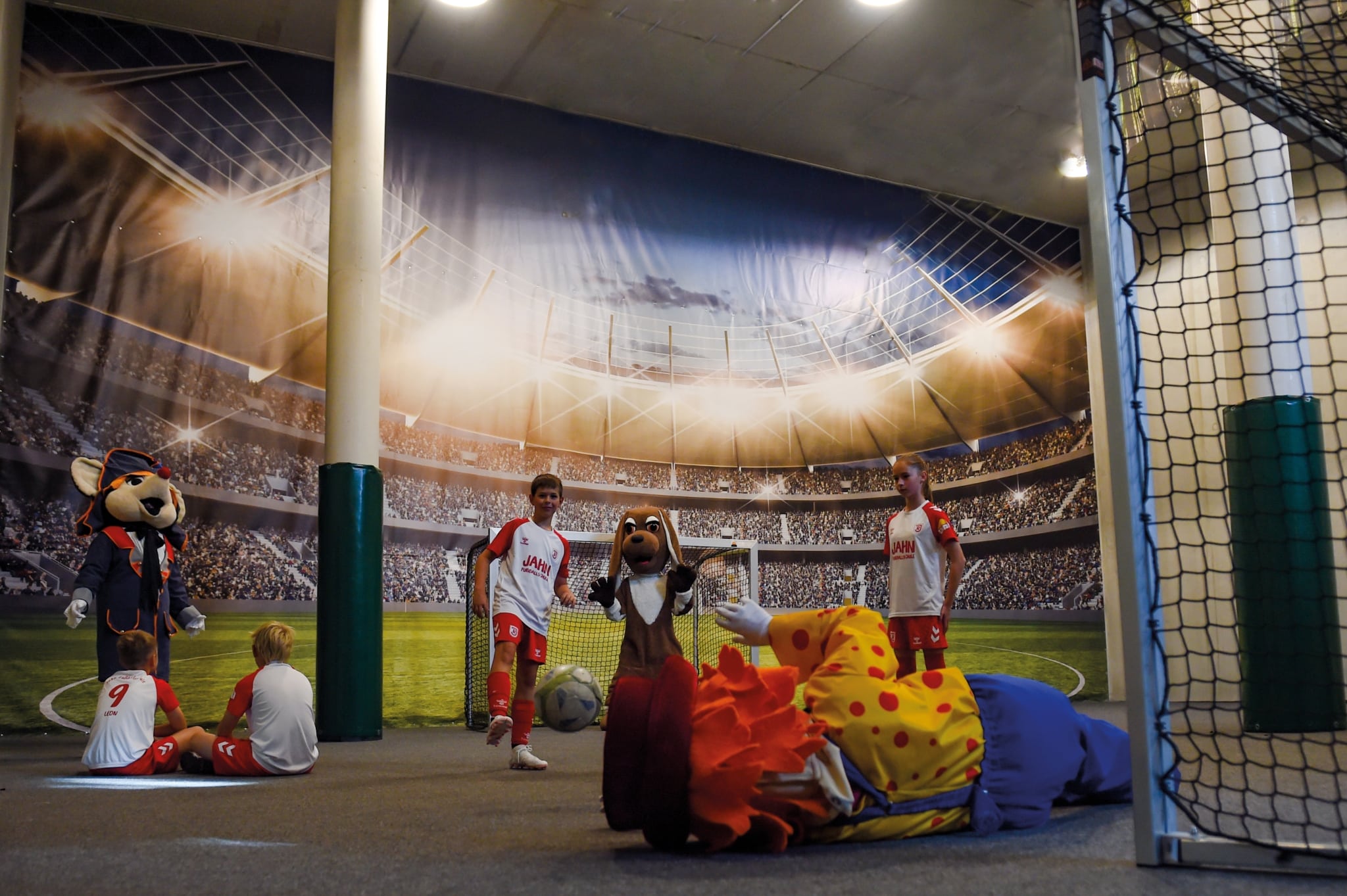 Indoor-Sporthalle mit Fußballmotiv: Kinder spielen vor einem Tor Fußball, begleitet von zwei fröhlichen Maskottchen in Kostümen – Stadionkulisse im Hintergrund sorgt für echtes Arena-Feeling.