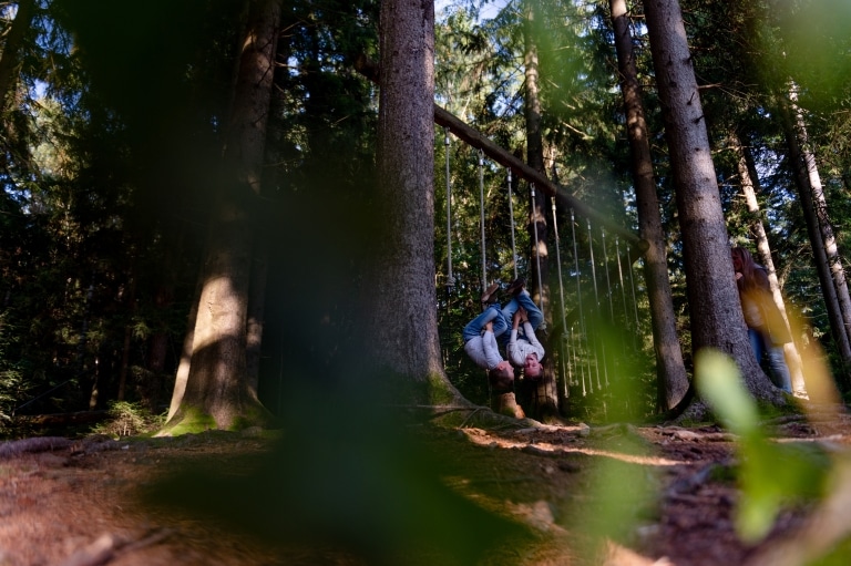 Zwei Jungen hängen kopfüber an Seilen auf einem Waldspielplatz im ULRICHSHOF, umgeben von hohen Bäumen und natürlicher Waldkulisse