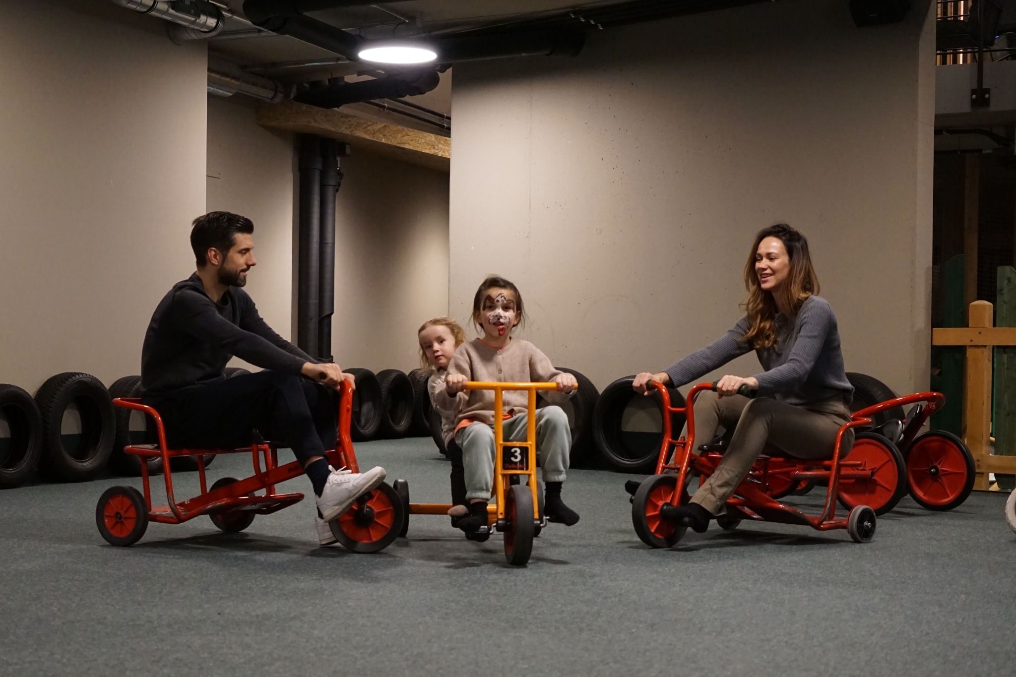 Familie mit zwei Erwachsenen und zwei Kindern auf roten Gokarts in einer Indoor-Spielhalle, ein Kind mit geschminktem Gesicht sitzt vorne auf einem Dreirad.
