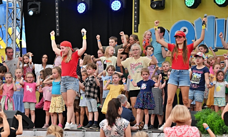 Eine große Gruppe von Kindern steht gemeinsam auf einer Bühne und macht mit erhobenen Armen eine Tanzbewegung. Zwei Animateurinnen in roten Shirts und Caps leiten die Kinder an und machen die Bewegungen vor. Im Hintergrund sind Bühnentechnik und Publikum zu sehen, die Szene wirkt wie ein lebendiger Programmpunkt bei einem Kinder-Event.