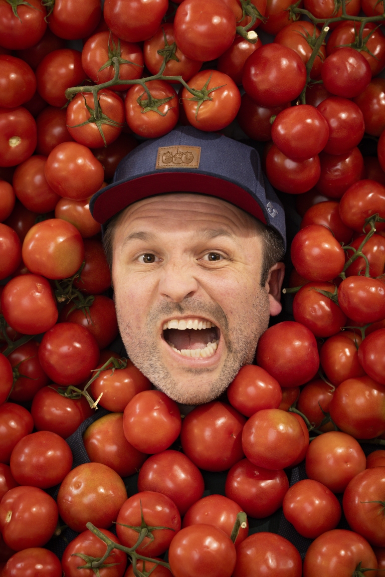 Ein Mann mit Baseballkappe schaut mit weit geöffnetem Mund und einem lebhaften Gesichtsausdruck in die Kamera. Sein Kopf ist dicht umgeben von vielen roten Tomaten, die das gesamte Bild ausfüllen.