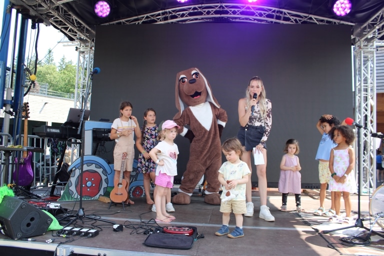 Mehrere Kinder stehen gemeinsam mit einer Moderatorin und einem Maskottchen in Hunde-Kostüm auf einer Bühne. Die Moderatorin hält ein Mikrofon und spricht ins Publikum, während die Kinder unterschiedlich aufmerksam oder verspielt reagieren. Im Hintergrund sind Musikinstrumente und Bühnentechnik zu sehen, die Szene wirkt wie ein Programmpunkt bei einem Kinderfestival.