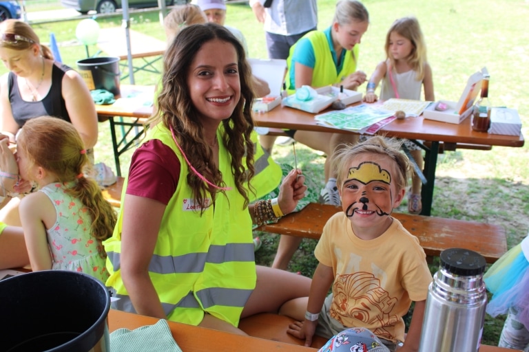 Eine Frau in einer gelben Warnweste sitzt an einem Tisch im Freien und lächelt in die Kamera, während sie einem Kind das Gesicht bemalt. Das Kind trägt ein Löwengesicht und grinst stolz. Im Hintergrund sitzen weitere Kinder an Tischen und basteln oder werden geschminkt, was auf eine lebendige Kinderbetreuungs- oder Festivalstation hinweist.