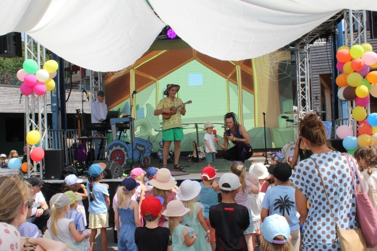 Eine Kindergruppe steht vor einer Bühne im Freien und schaut aufmerksam zu. Auf der Bühne spielen Musiker, während ein Entertainer sich zu einem kleinen Kind hinunterbeugt und ihm ein Mikrofon hinhält. Bunte Luftballons und eine fröhliche Bühnenkulisse sorgen für eine lebendige, familienfreundliche Atmosphäre.